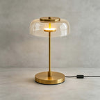 Clérachic table lamp with a clear glass dome lit against a grey wall, the brass cap and glowing ring visible under the glass.