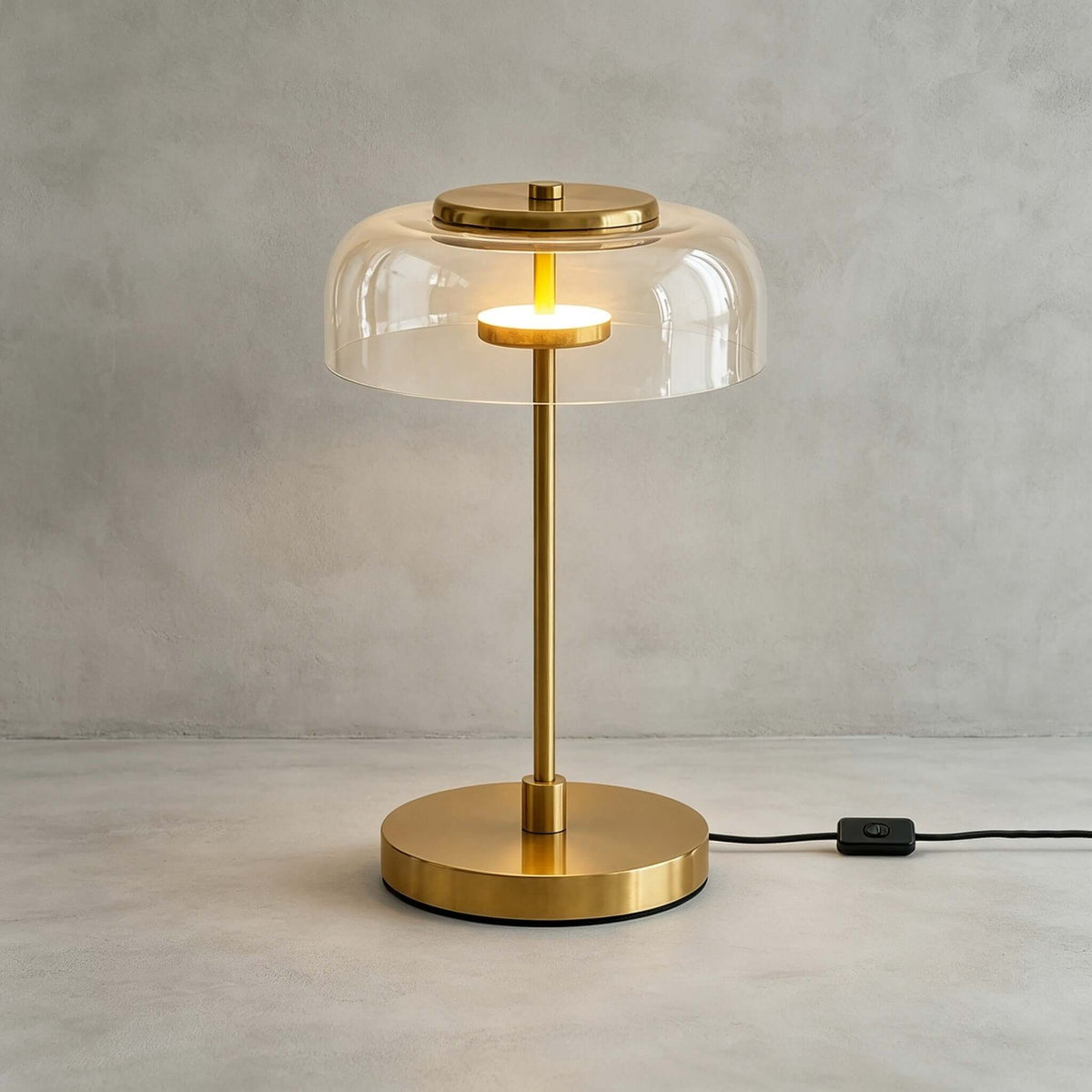 Clérachic table lamp with a clear glass dome lit against a grey wall, the brass cap and glowing ring visible under the glass.