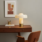 A Clérachic cream table lamp with a brass base glows on a walnut desk, beside a brass pen pot and a stack of books.