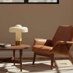 Beside a round walnut coffee table and a tan leather armchair, this Clérachic cream and brass lamp stands in warm afternoon light.
