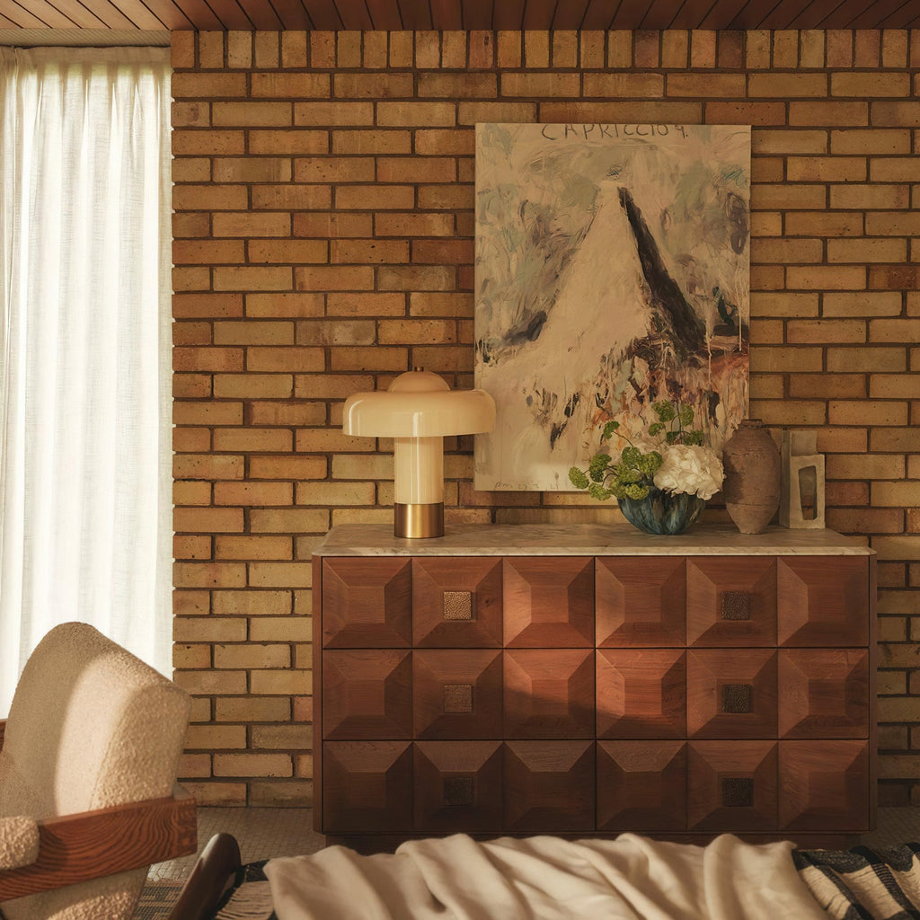 The Clérachic cream and brass lamp on a carved walnut sideboard, in front of a brick wall and an abstract painting.