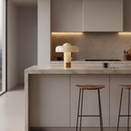 A small Clérachic cream lamp with a brass base on a marble kitchen island, with wooden bar stools below.