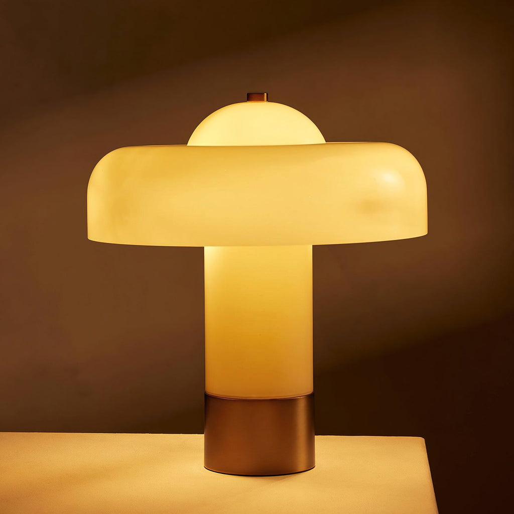 Lit Clérachic table lamp glowing amber through its cream glass stem and shade, on a pale surface against a dark brown backdrop.
