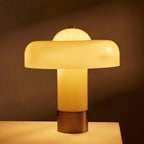 Lit Clérachic table lamp glowing amber through its cream glass stem and shade, on a pale surface against a dark brown backdrop.