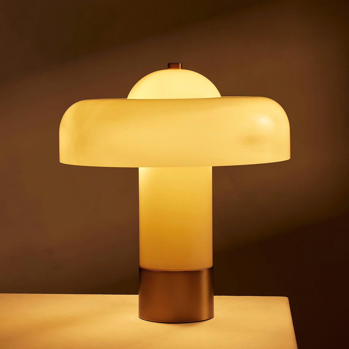 Lit Clérachic table lamp glowing amber through its cream glass stem and shade, on a pale surface against a dark brown backdrop.