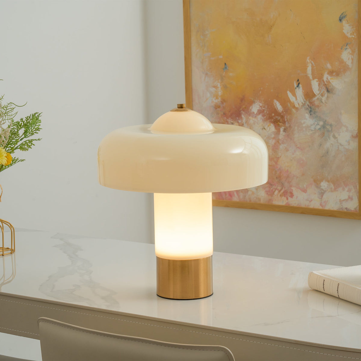 The stem of this Clérachic table lamp glows warm white under its cream shade, on a white marble desk by a golden painting.
