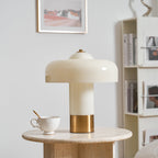 A Clérachic cream table lamp with a brass base, on a travertine round table beside a lace-textured teacup and a bookcase.