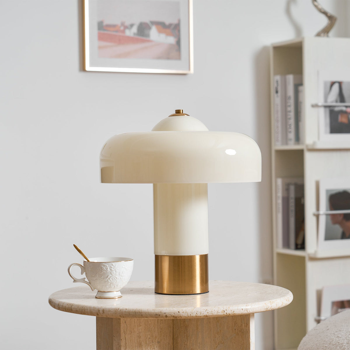 A Clérachic cream table lamp with a brass base, on a travertine round table beside a lace-textured teacup and a bookcase.