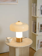 Lit Clérachic cream lamp on a travertine round table, its stem glowing white, beside a teacup and a shelf of books.