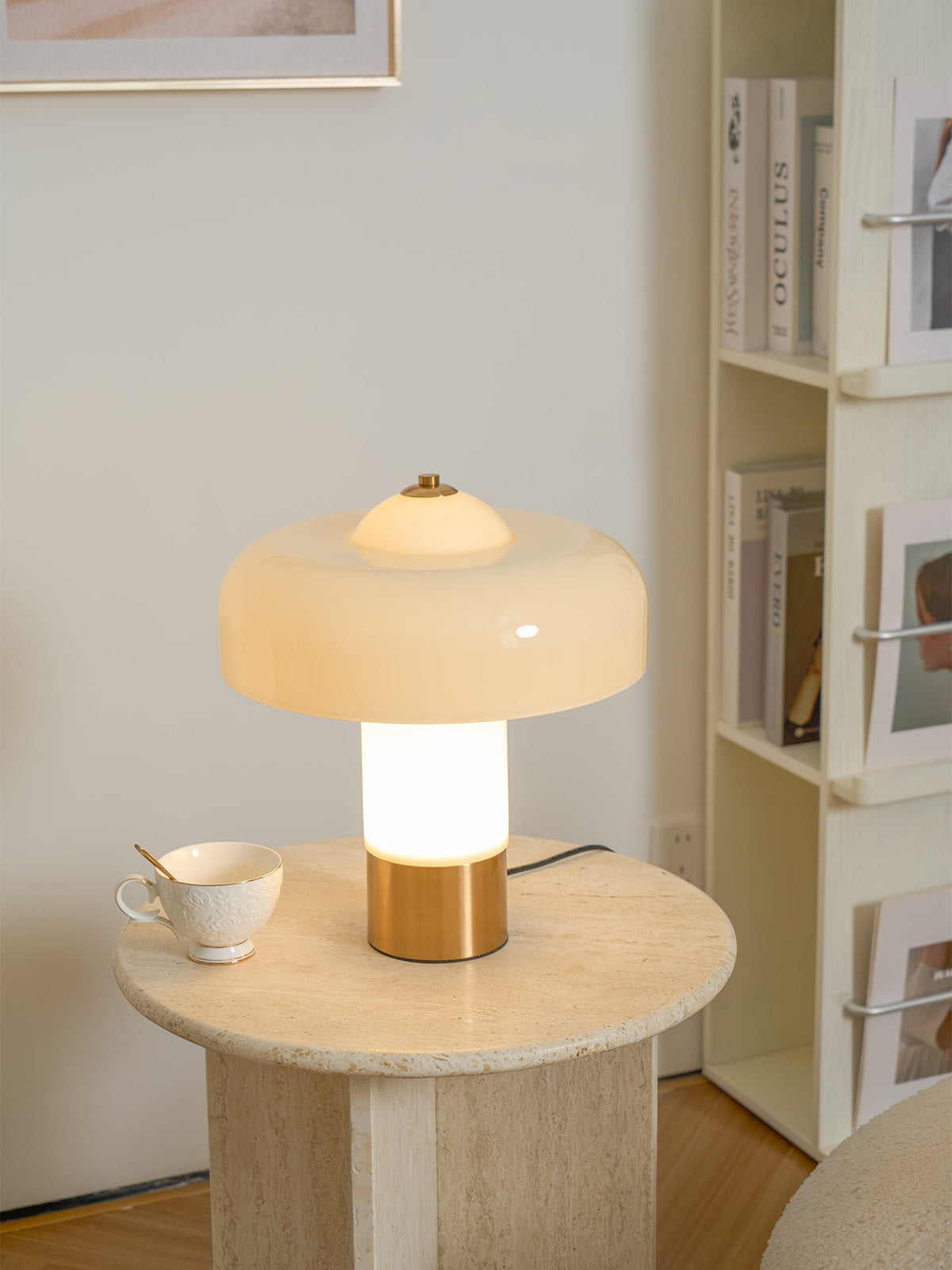 Lit Clérachic cream lamp on a travertine round table, its stem glowing white, beside a teacup and a shelf of books.