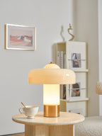 A Clérachic cream table lamp lit on a travertine side table, with a framed landscape print and a bookcase behind.