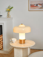 Clérachic cream and brass lamp, lit, on a round stone table beside a white fireplace and a stack of logs.