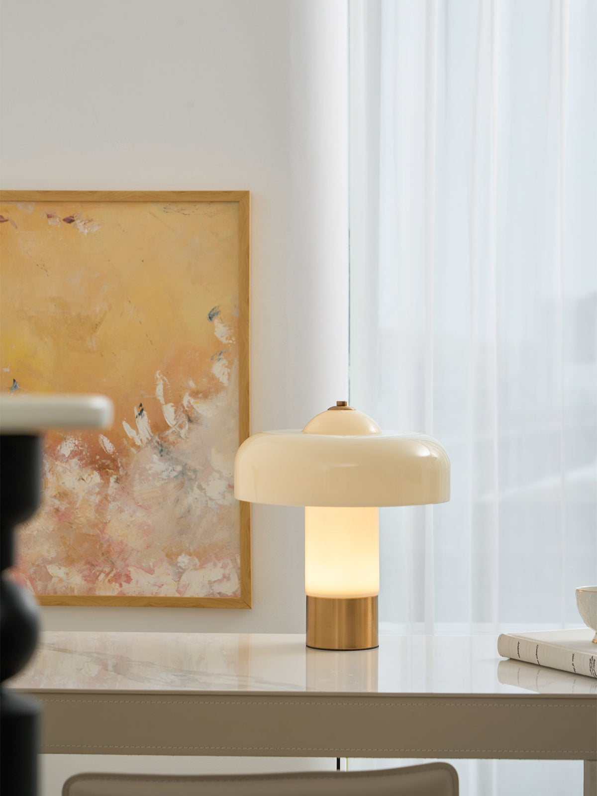 A Clérachic cream table lamp glows on a white desk in front of a golden abstract painting and sheer curtains.
