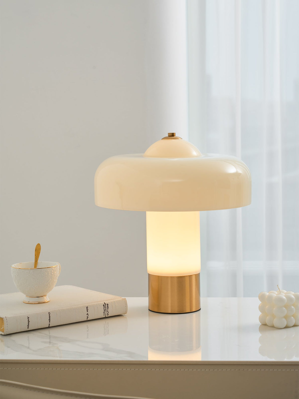 The Clérachic table lamp lit on a white marble desk with a teacup on a book and a small bobbled candle beside it.