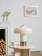 Unlit Clérachic cream lamp with a brass base on a round travertine table, by a white fireplace and framed print.