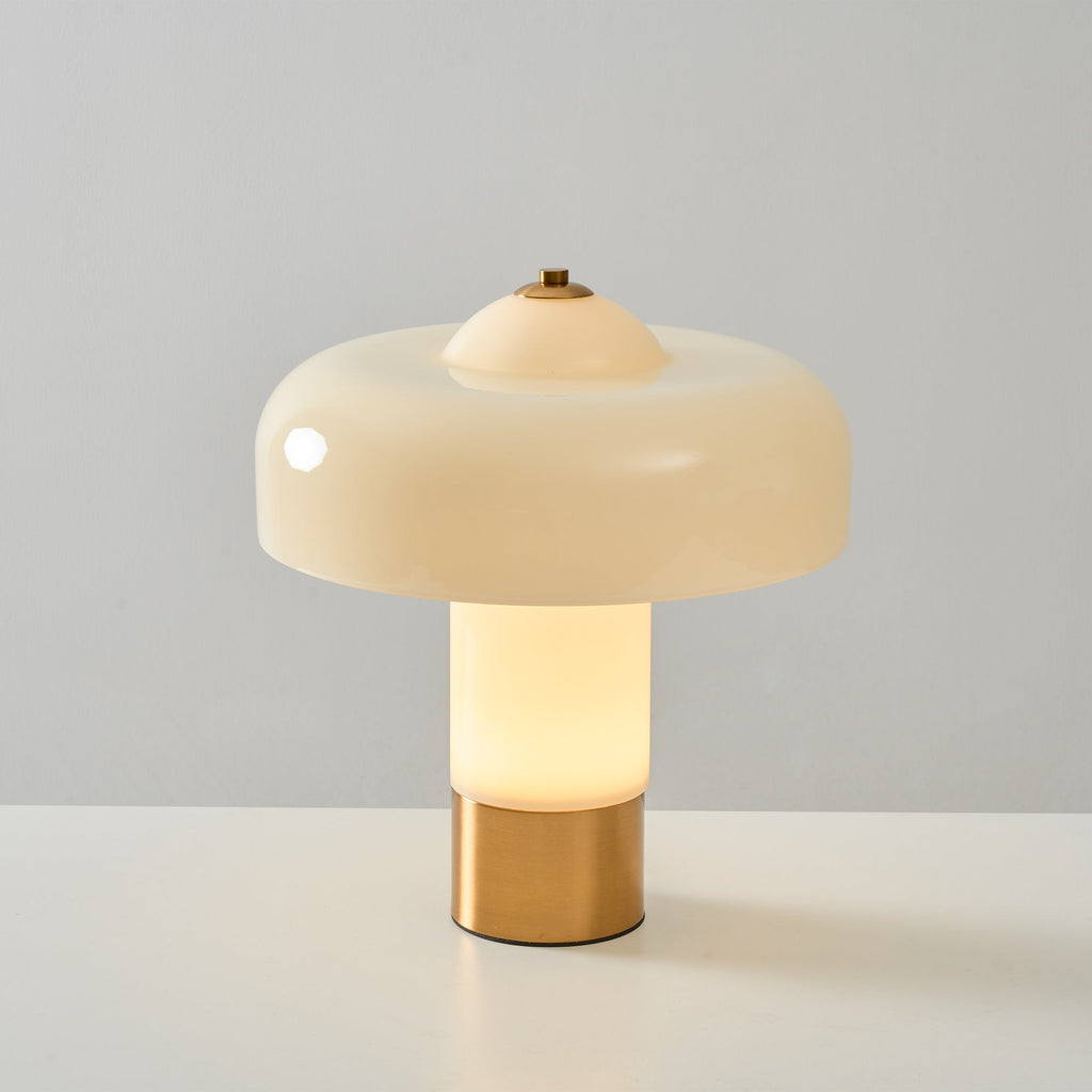 A Clérachic table lamp lit from inside, its cream stem glowing white above a brass base, against a grey wall.