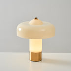 A Clérachic table lamp lit from inside, its cream stem glowing white above a brass base, against a grey wall.