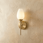 Clérachic brass wall lamp with a tulip-shaped frosted glass shade on a slim stem and round backplate, glowing warm on a plaster wall.