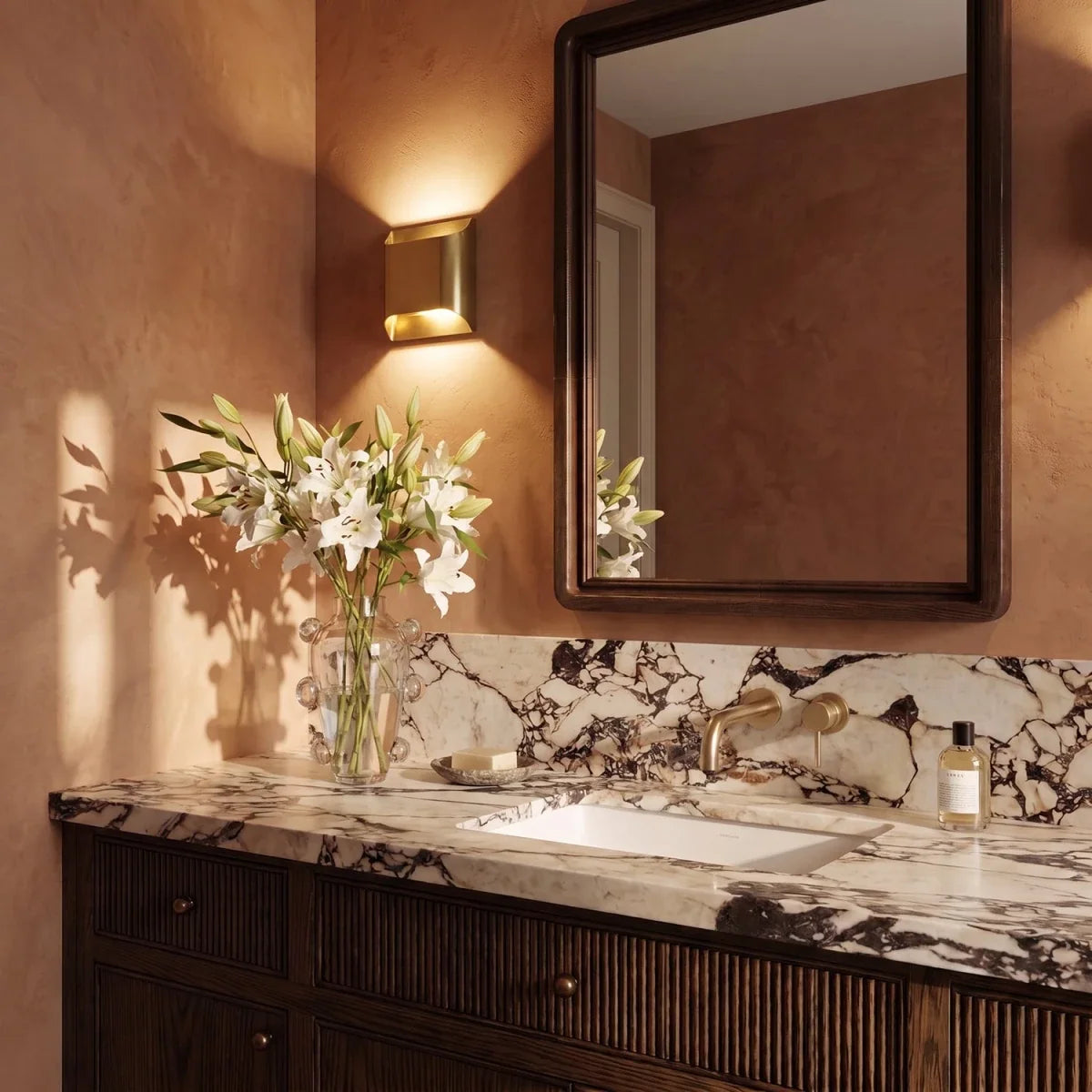 Clérachic square brass wall light beside a mirror above a marble vanity in a warm terracotta-toned bathroom.