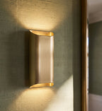 Clérachic tall curved brushed brass wall light close up on green grasscloth, glowing warm from top and bottom.