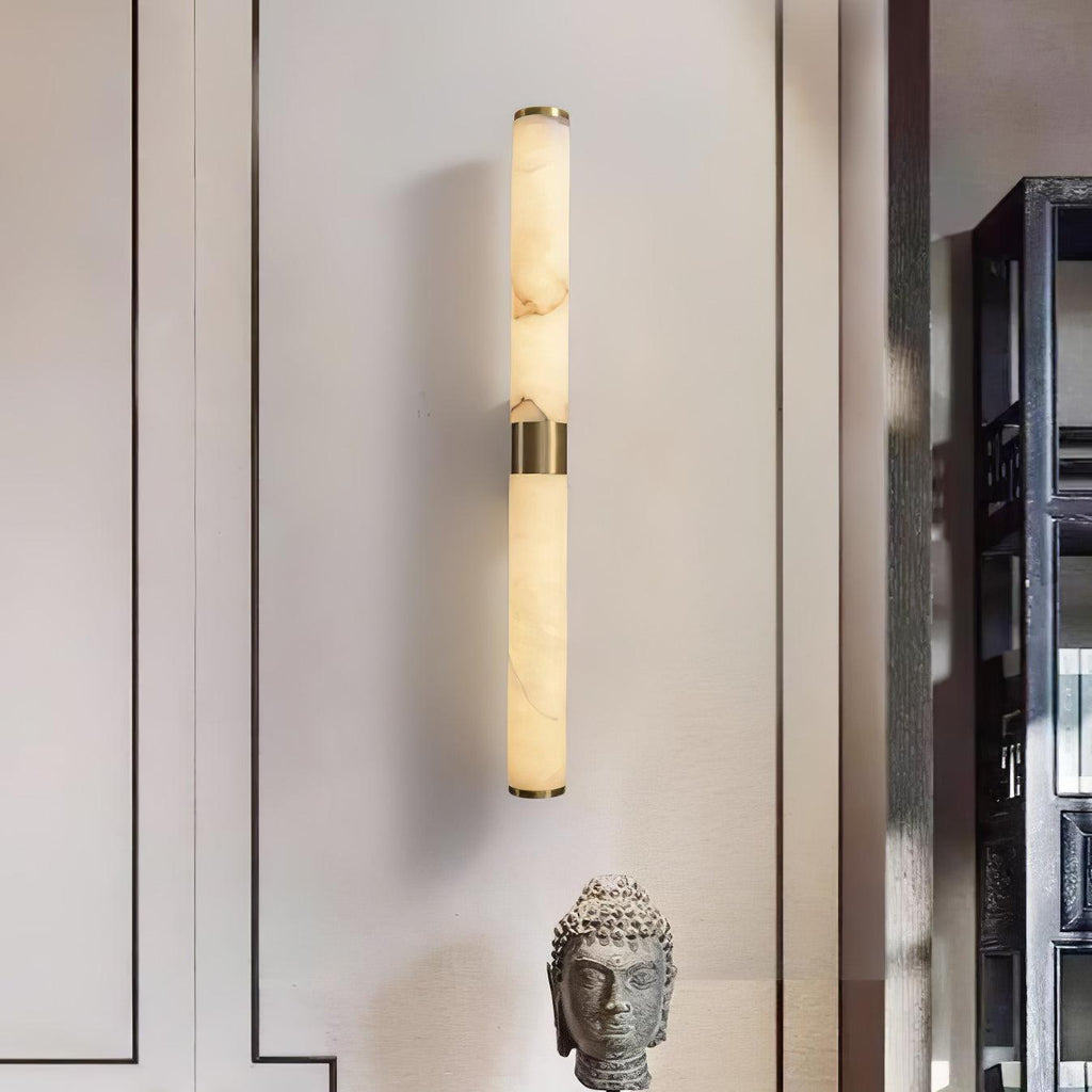 A slim brass-banded alabaster tube wall light glows softly on a pale wall, beside a stone head sculpture, from Clérachic.