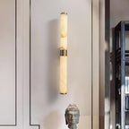 A slim brass-banded alabaster tube wall light glows softly on a pale wall, beside a stone head sculpture, from Clérachic.