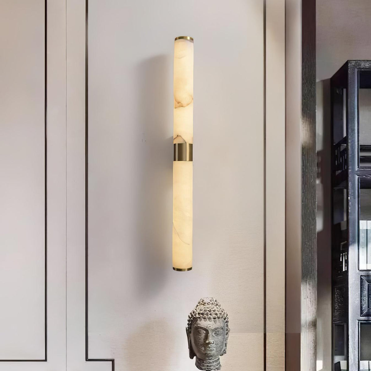 A slim brass-banded alabaster tube wall light glows softly on a pale wall, beside a stone head sculpture, from Clérachic.