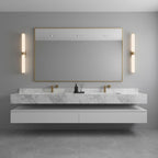 Either side of a gold-framed mirror over a marble basin shelf, a pair of alabaster tube lights glow warm, from Clérachic.