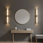 A pair of alabaster tube wall lights with brass bands, one on each side of a round mirror above a pale wood console, from Clérachic.