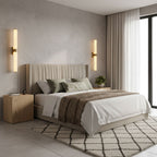 On the wall behind a bed with a woven throw and a patterned rug, two glowing alabaster tubes from Clérachic stand either side.