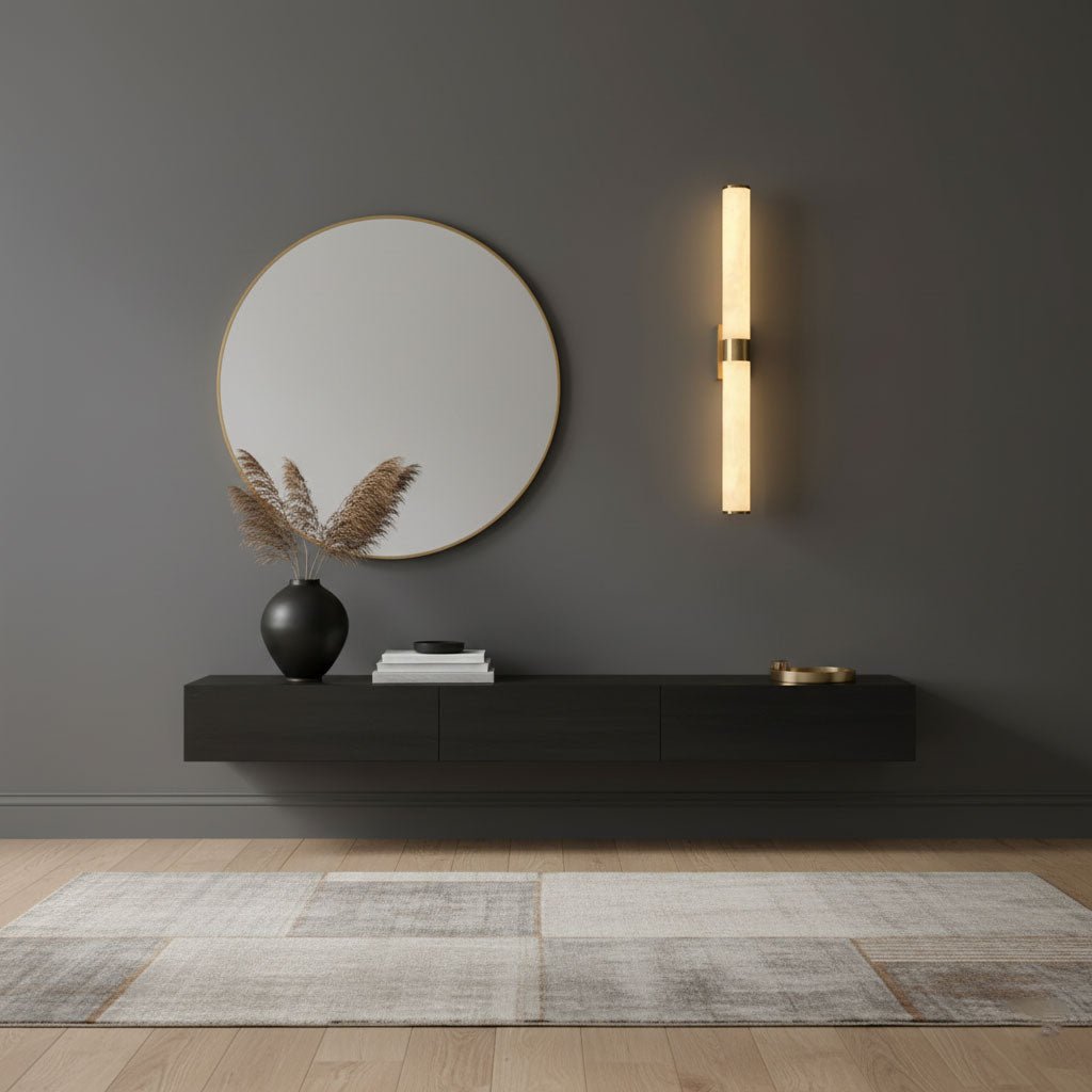 By a round mirror and a floating black shelf on a grey wall, a Clérachic alabaster tube wall light glows from a brass band.