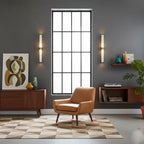 Two alabaster tube wall lights on a grey wall flank a tall paned window, above a mid-century sideboard and tan armchair, from Clérachic.
