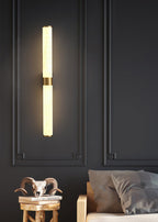 A lit alabaster tube wall light on a dark panelled wall, with a brass band at the centre and a bedroom corner below, from Clérachic.