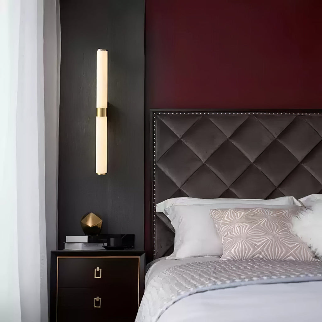 By a velvet quilted headboard with a deep red wall behind, an alabaster tube wall light glows above a dark bedside chest, from Clérachic.