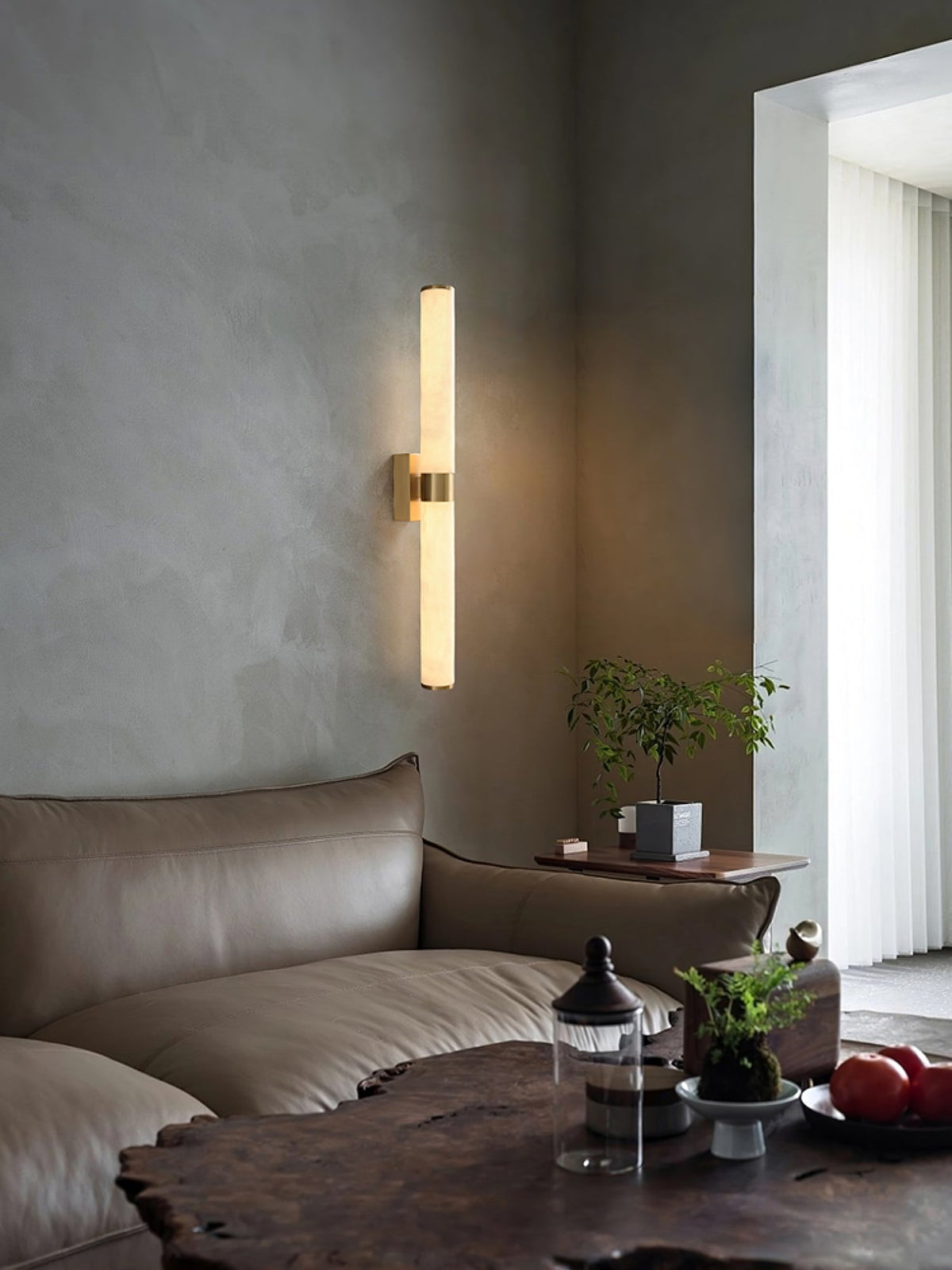 Above a taupe leather sofa and a live-edge wood table, a slim alabaster tube light glows on a plaster-grey wall, from Clérachic.