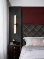 A wider view of this Clérachic alabaster tube light above a dark bedside chest, with a quilted headboard and a red wall behind it.