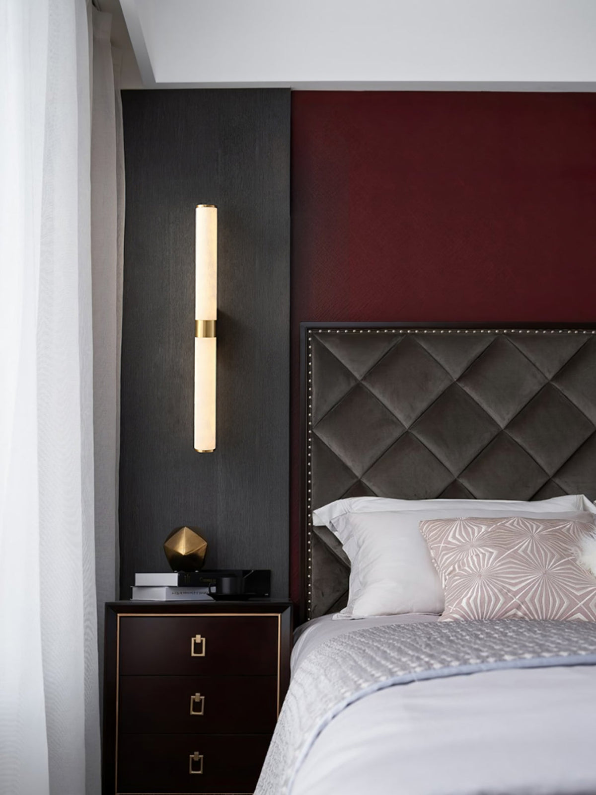 A wider view of this Clérachic alabaster tube light above a dark bedside chest, with a quilted headboard and a red wall behind it.