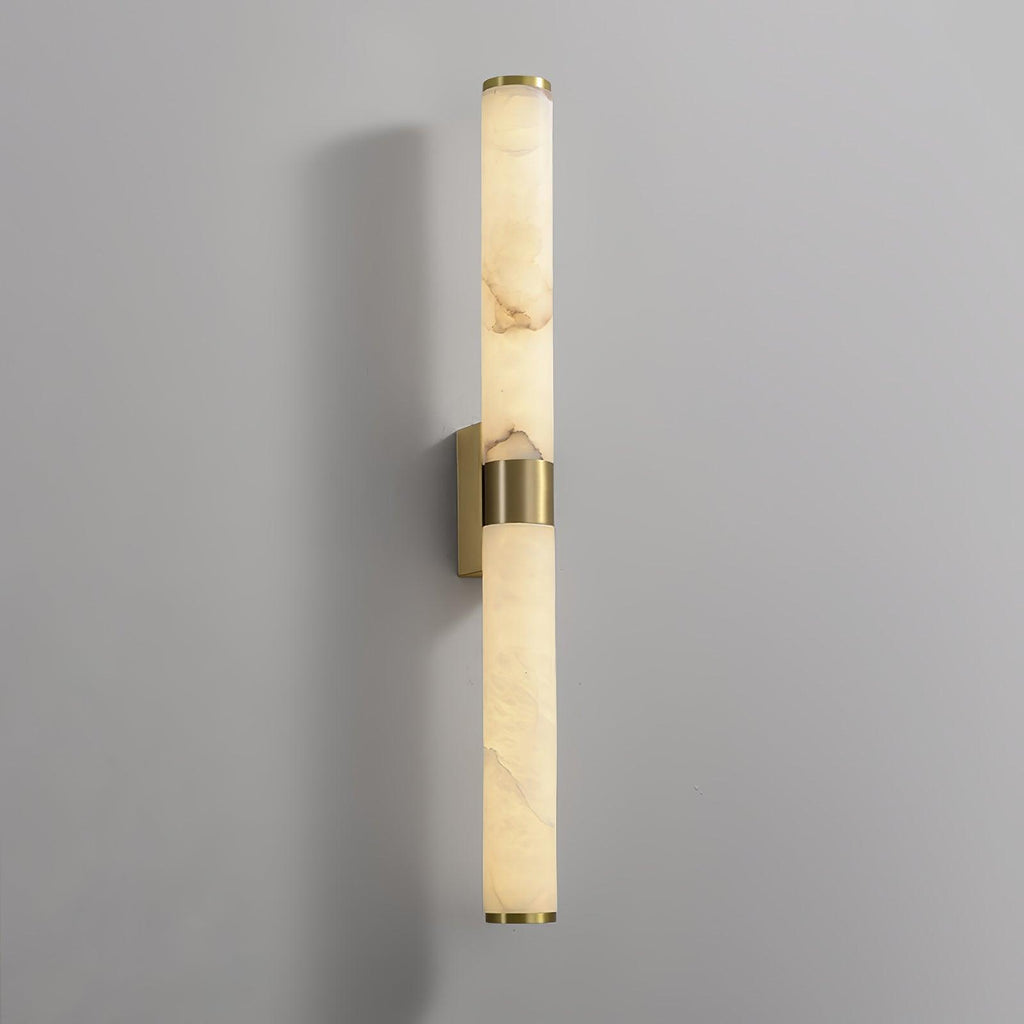 A tall alabaster tube with a brass band on a grey wall, with its brass back plate showing behind the band, from Clérachic.