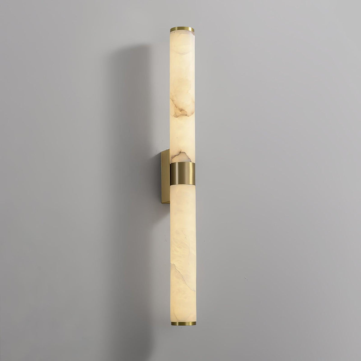 A tall alabaster tube with a brass band on a grey wall, with its brass back plate showing behind the band, from Clérachic.
