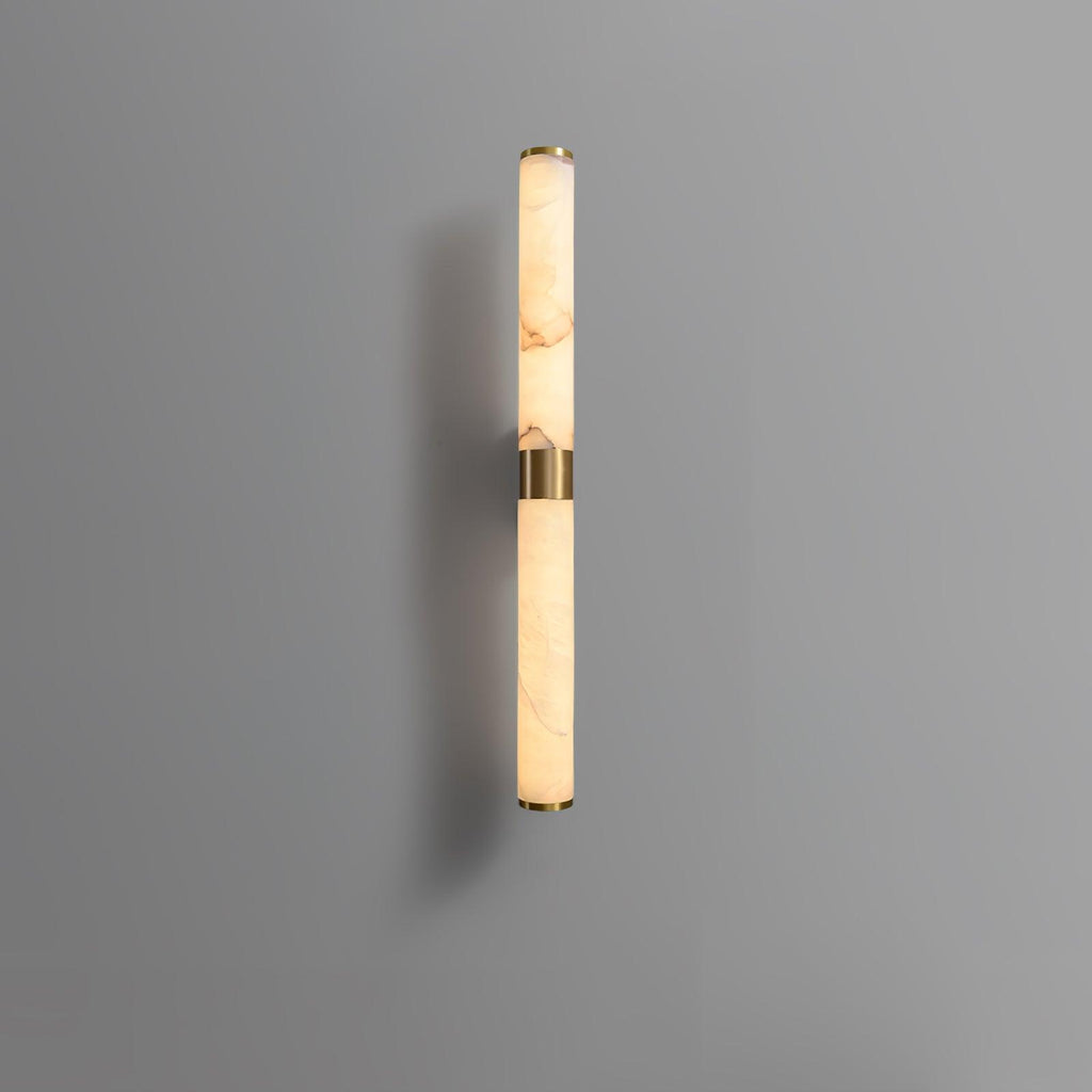 A shorter alabaster and brass tube wall light on a grey wall, with its brass band at the middle, from Clérachic.