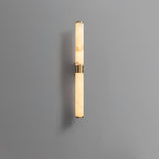 A shorter alabaster and brass tube wall light on a grey wall, with its brass band at the middle, from Clérachic.