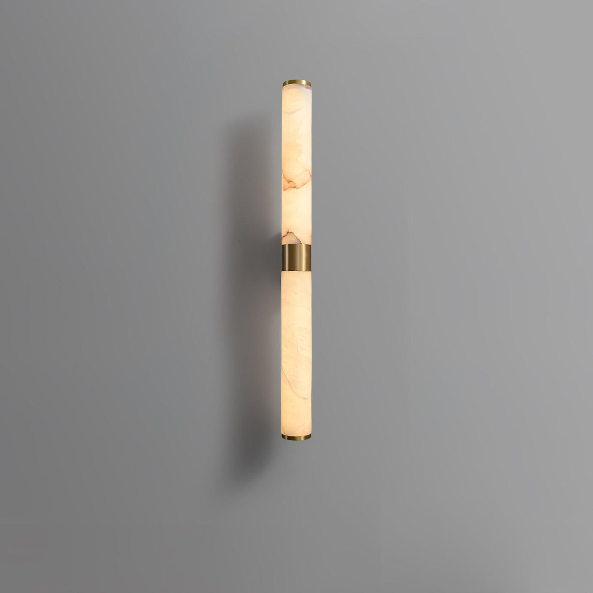 A shorter alabaster and brass tube wall light on a grey wall, with its brass band at the middle, from Clérachic.