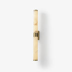 A veined alabaster tube wall light with a brass band and plate, on a soft white background with a faint shadow, from Clérachic.