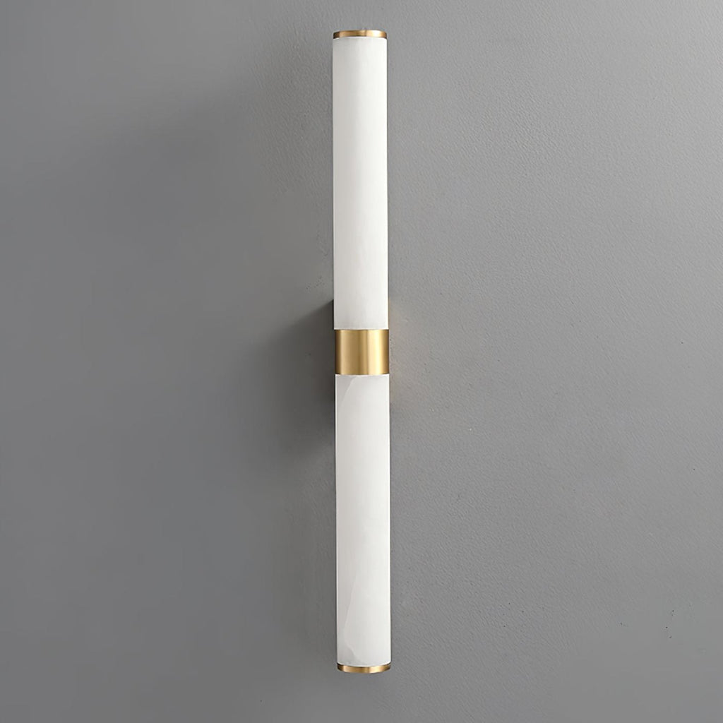 A pale white alabaster tube wall light with a brass band, on a grey wall, with little visible veining, from Clérachic.