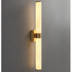 Lit warm, a Clérachic wall light with two alabaster tubes and a brass band and plate casts amber light up and down a grey wall.