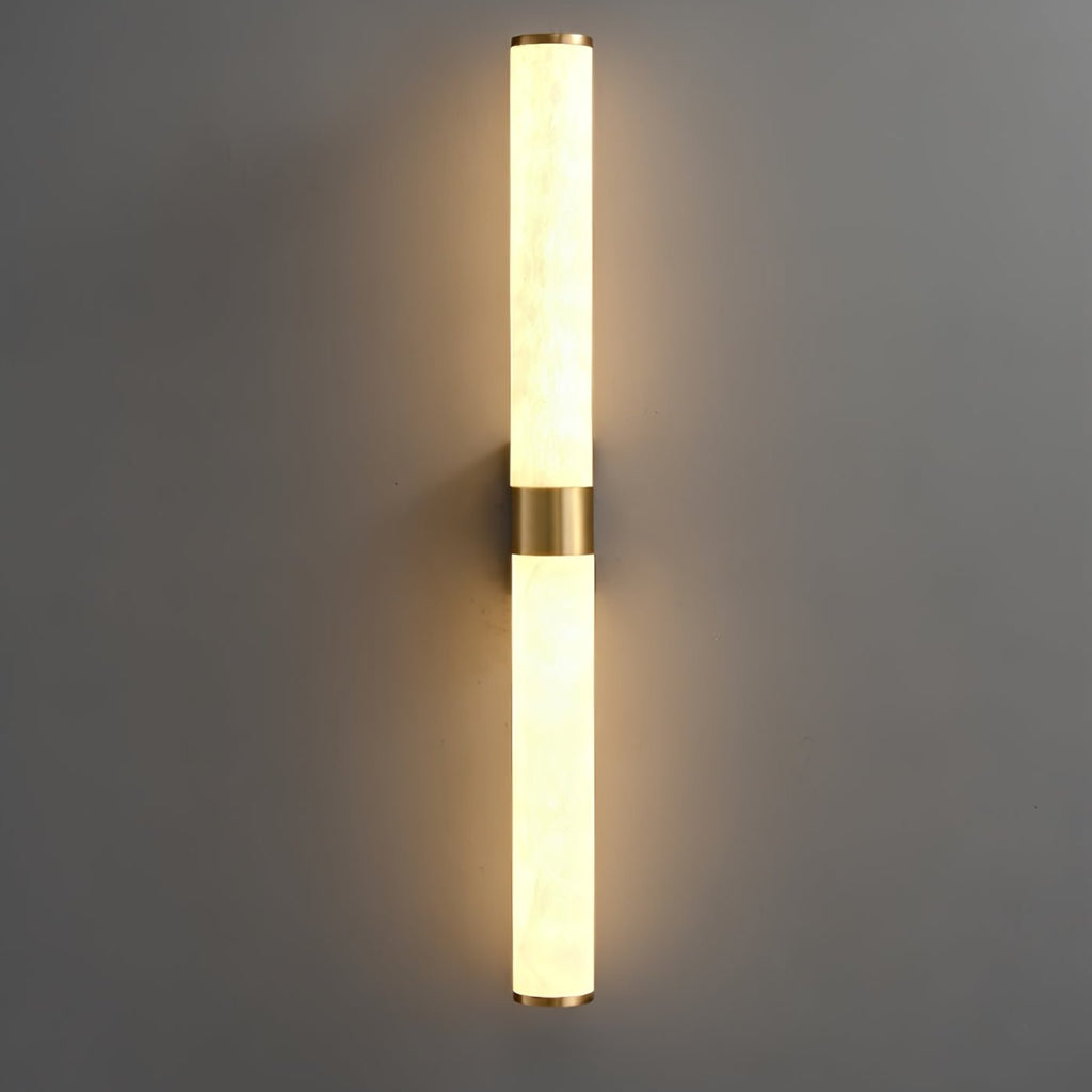 Glowing alabaster tubes with a brass band at the centre cast a warm halo on a dark grey wall, from Clérachic.