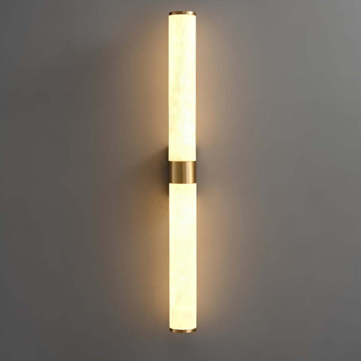 Glowing alabaster tubes with a brass band at the centre cast a warm halo on a dark grey wall, from Clérachic.
