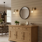 Two Clérachic brass wall lights with clear glass shades flank a round black-framed mirror above a light oak sideboard.