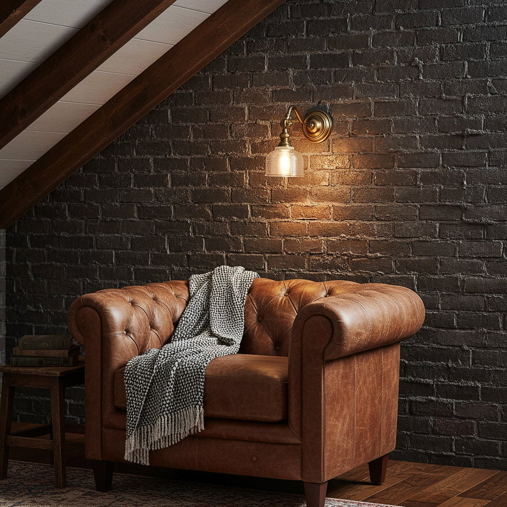 A Clérachic brass wall light with a small glass shade glows against a dark brick wall, above a tan leather armchair.