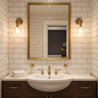 Two Clérachic wall lights with glass shades flank a gold-framed mirror on a white tiled wall, above a basin with brass taps.